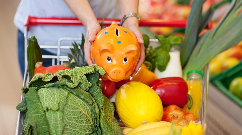 Woman Keeping Piggybank In Shopping Cart Full Of Groceries